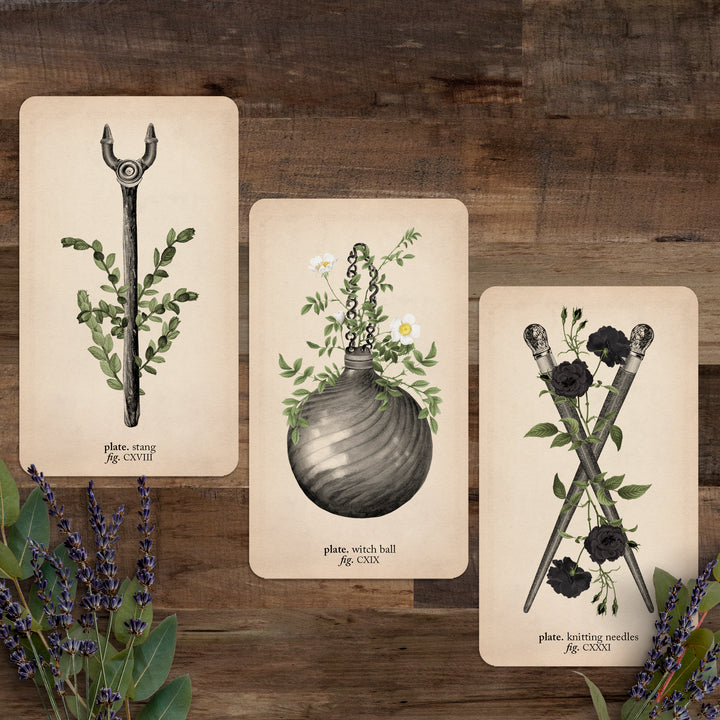 Three cards (stang, witch ball, and knitting needles) against a wooden background with lavender flowers below